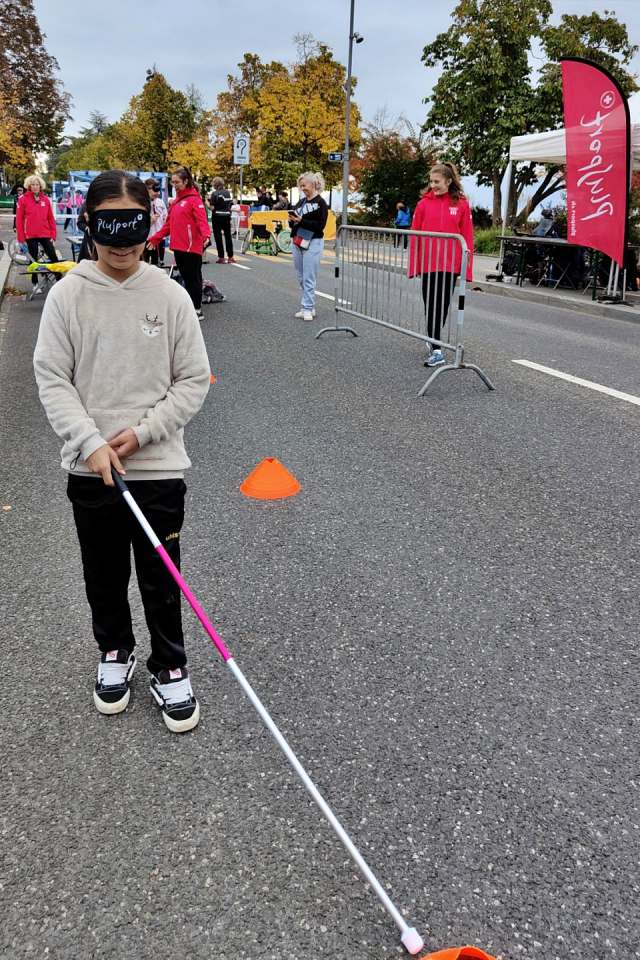 Ein Kind mit einer Augenbinde und einem weißen Stock navigiert über einen mit orangefarbenen Kegeln markierten Weg auf einer Stadtstraße, während im Hintergrund Menschen und Veranstaltungszelte zu sehen sind.