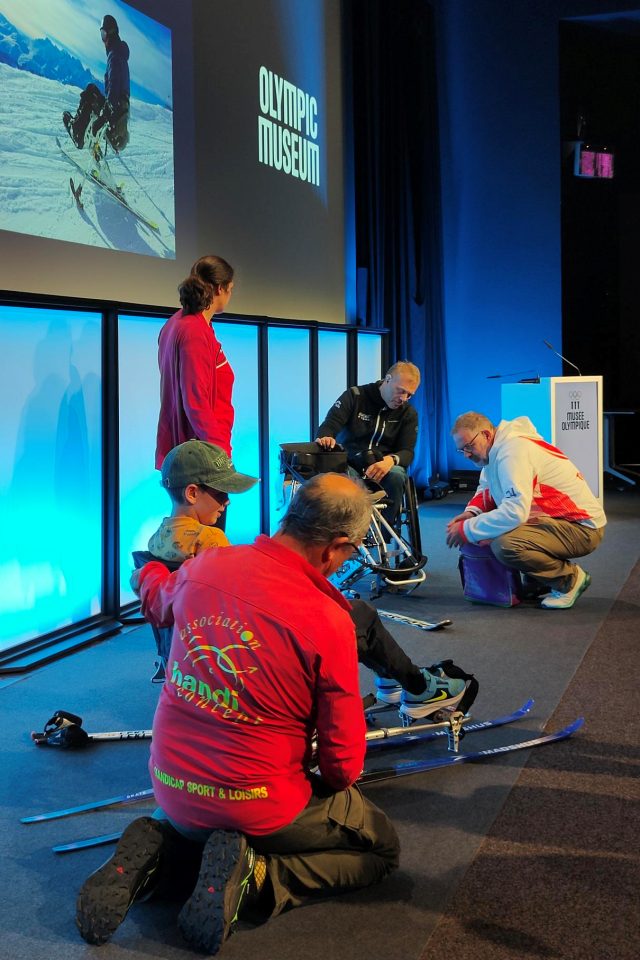 Mehrere Personen versammeln sich im Olympischen Museum, eine Person im Rollstuhl und andere, die mit der Skiausrüstung helfen; ein großes Foto eines Sitzskis wird an die Wand projiziert.