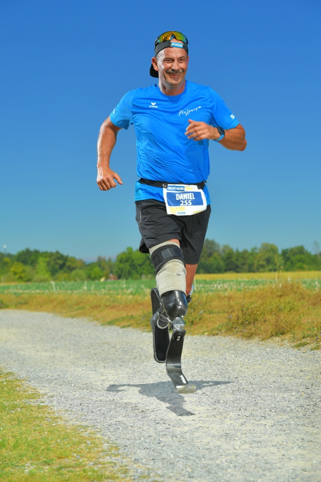 Ein männlicher Läufer mit einer Beinprothese, der ein blaues Hemd, schwarze Shorts und eine Mütze trägt, läuft auf einem Kiesweg im Freien unter einem klaren blauen Himmel. Er lächelt und scheint voller Energie zu sein.