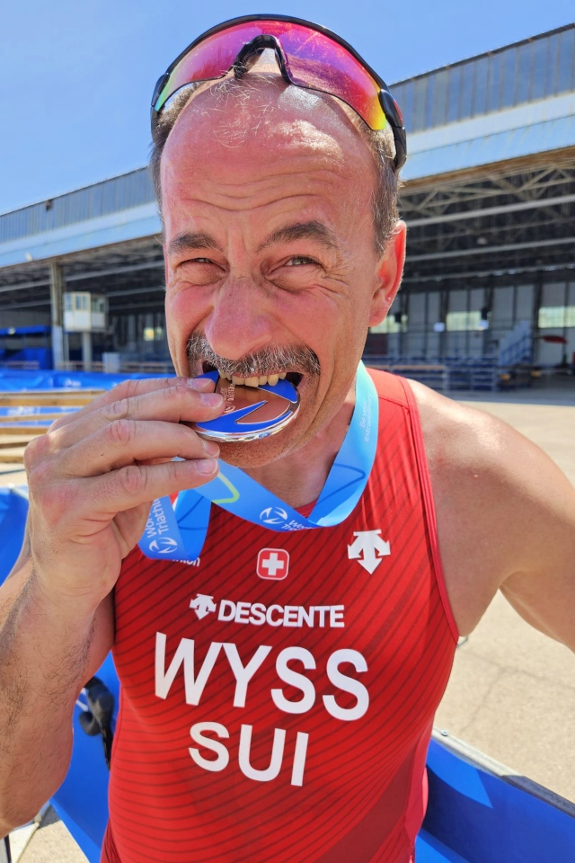 Ein männlicher Athlet in einem roten Schweizer Triathlonanzug mit dem Aufdruck WYSS SUI beißt lächelnd in eine Medaille. Er steht im Freien vor einem großen Gebäude und trägt eine Sonnenbrille auf dem Kopf.
