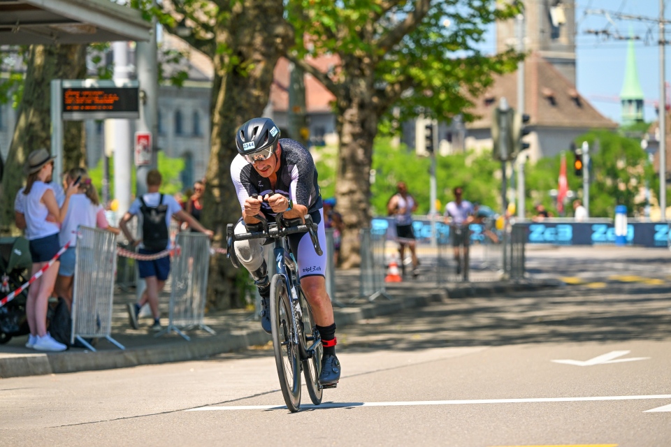 Ein Radfahrer in Rennkleidung fährt während eines Wettkampfs auf einer städtischen Straße Fahrrad. Die Zuschauer stehen hinter Absperrungen, und im Hintergrund sind an einem sonnigen Tag Bäume und historische Gebäude zu sehen.