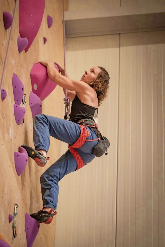Eine Person mit lockigem Haar, die einen Klettergurt, Jeans und Kletterschuhe trägt, erklimmt eine Indoor-Kletterwand mit großen lila Griffen. Sie greift mit einem Arm nach oben und sieht konzentriert und entschlossen aus.