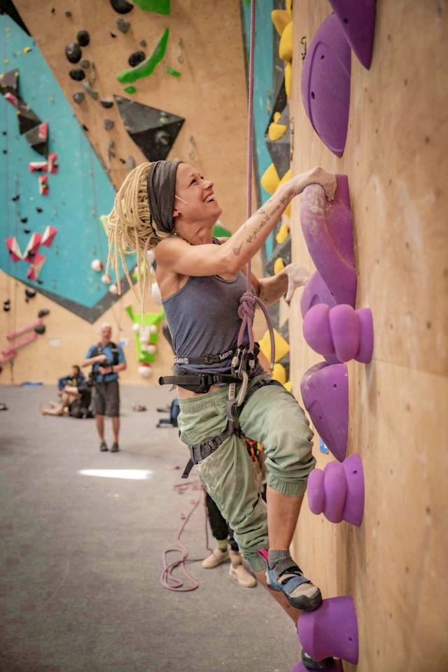 Eine Frau mit blonden Dreadlocks und einem Stirnband klettert lächelnd an einer Indoor-Felswand mit großen lilafarbenen Griffen empor. Sie trägt eine grüne Hose, ein blaues Tank-Top und eine Kletterausrüstung. Im Hintergrund sind andere Personen zu sehen.
