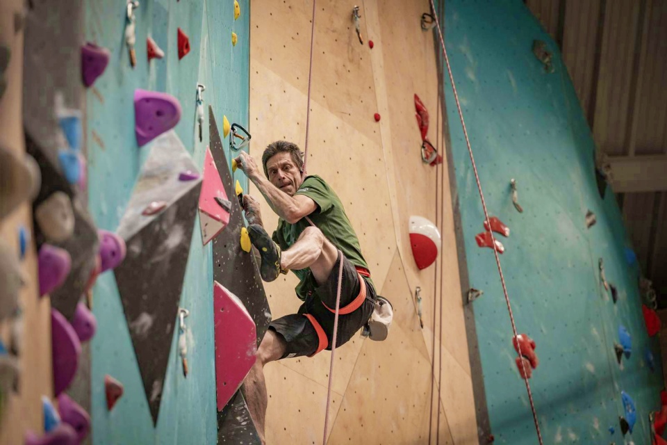 Ein Mann mit Klettergurt und grünem Hemd klettert an einer bunten Indoor-Kletterwand und hält sich mit Händen und Füßen fest, während er in Sicherungsseile eingehängt ist.