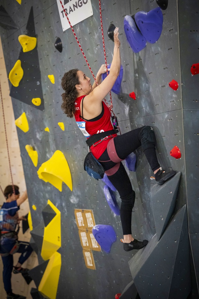 Eine Frau in rotem Shirt und schwarzen Leggings klettert während eines Wettkampfs eine Indoor-Kletterwand mit violetten und gelben Griffen. Sie greift nach einem roten Seil und konzentriert sich auf ihren nächsten Zug. Im Hintergrund ist eine weitere Kletterin zu sehen.