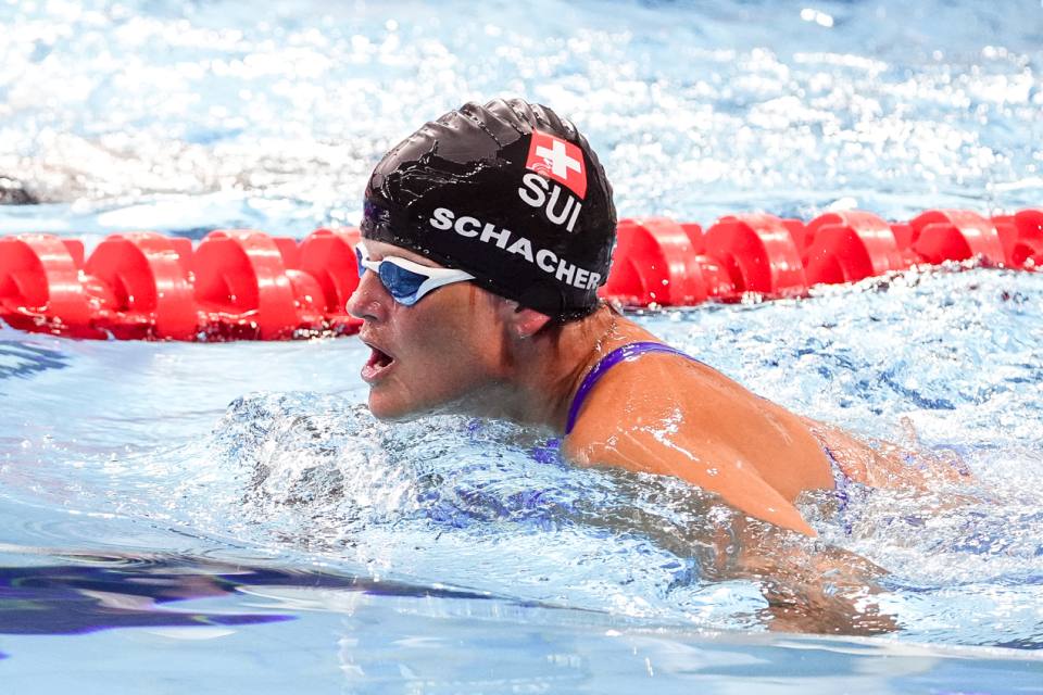 Ein Schwimmer, der eine schwarze Badekappe mit der Aufschrift SUI und eine Schwimmbrille trägt, tritt in einem Schwimmbecken in der Nähe einer roten Bahntrennung mit offenem Mund an, möglicherweise während eines Atemzugs.