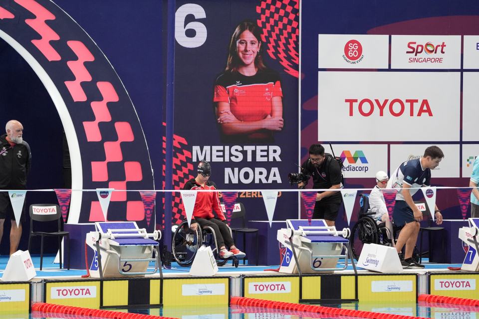 Mehrere Sportler in Rollstühlen bereiten sich in einem Hallenbad auf einen Schwimmwettkampf vor. Hinter Bahn 6 hängt ein großes Banner mit der Aufschrift "Meister Nora" und ihrem Foto. Im Hintergrund sind Sponsoren der Veranstaltung wie Toyota zu sehen.