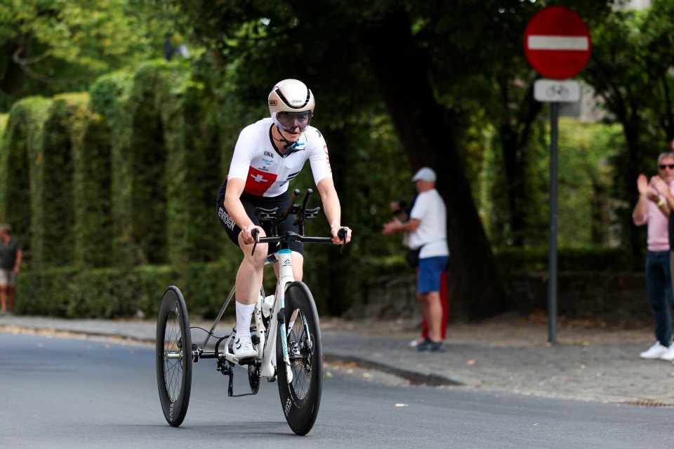 Ein Radfahrer, der einen weißen Helm und ein Schweizer Trikot trägt, fährt mit einem dreirädrigen Handbike auf einer Straße, mit Zuschauern und Grün im Hintergrund an einem sonnigen Tag.