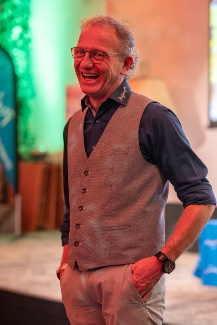 Ein lächelnder älterer Mann mit lockigem grauem Haar und Brille trägt eine graue Weste über einem dunklen Hemd und steht mit einer Hand in der Tasche in einem warm beleuchteten Innenraum.