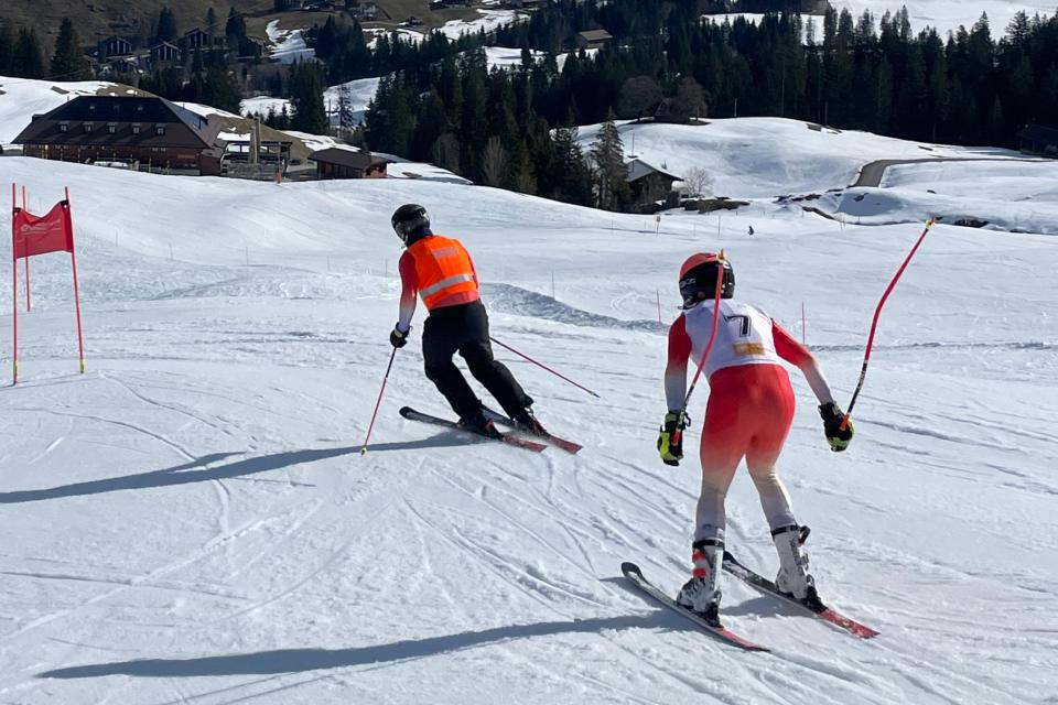 Zwei Skifahrer mit Helmen und Skianzügen rasen auf einer verschneiten Piste bergab, vorbei an roten Torflaggen. Im Hintergrund sind schneebedeckte Hügel, Bäume und Berghütten unter einem klaren Himmel zu sehen.