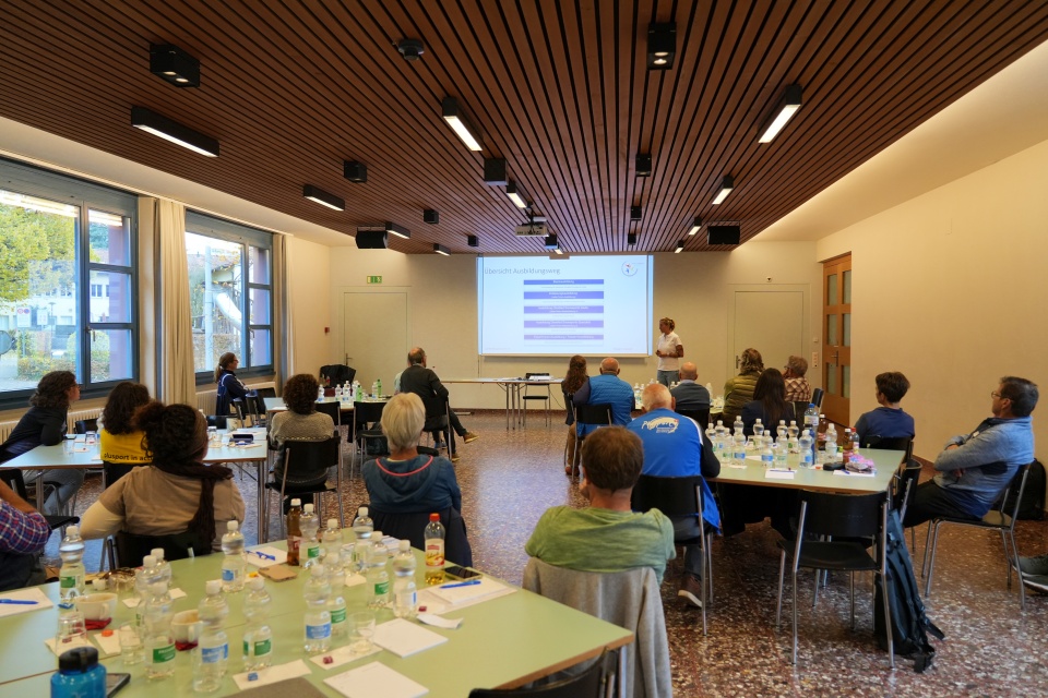 Eine Gruppe von Personen sitzt an Tischen in einem Konferenzraum und blickt auf einen Redner und eine Projektionsfläche, auf der ein Diagramm zu sehen ist. Auf den Tischen stehen Flaschen und Papiere, und große Fenster lassen von links natürliches Licht herein.