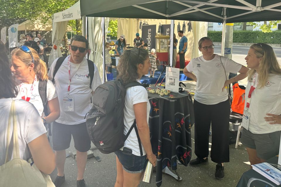 Eine Gruppe von Menschen steht und unterhält sich an einem Veranstaltungsstand im Freien unter einem Vordach. Einige tragen weiße Hemden mit Namensschildern. Auf dem Tisch liegen Broschüren und Gegenstände, und im Hintergrund sind andere Stände und Teilnehmer zu sehen.