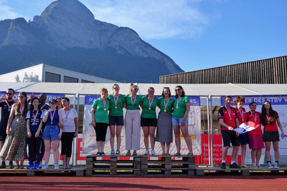 Drei Frauenmannschaften stehen mit Medaillen um den Hals auf dem Podium und feiern an einem sonnigen Tag vor einer Bergkulisse. Die Siegermannschaft in grünen Trikots steht in der Mitte, flankiert von den Teams auf den Plätzen zwei und drei.