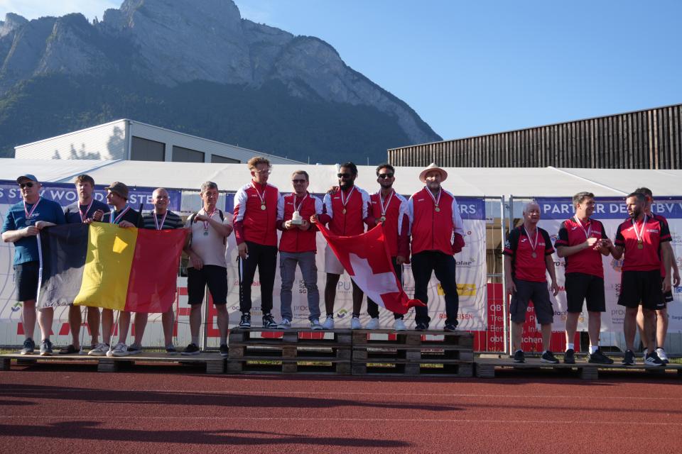 Sportler mit Medaillen stehen auf einem Podium und halten Nationalflaggen in den Händen, im Hintergrund ist ein Berg zu sehen. Neben ihnen stehen weitere Personen, von denen einige klatschen oder ihre Handys in der Hand halten – eine sonnige Sportveranstaltung im Freien.