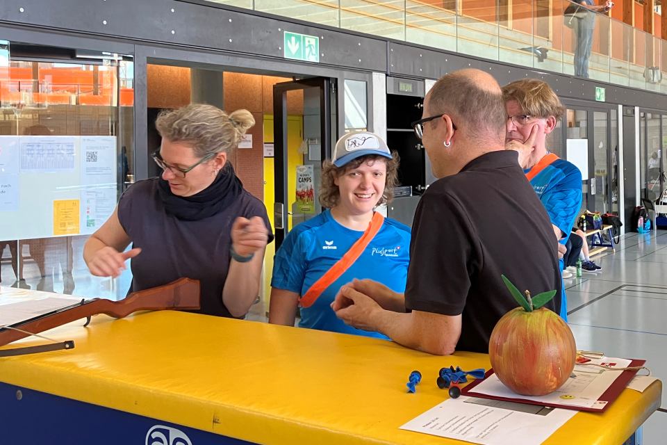 Vier Personen stehen an einem gelben Tisch in einer Turnhalle. Eine Person hantiert mit einem Holzgewehr, während eine andere in einem blauen Sporthemd und einer Mütze lächelt. Auf dem Tisch liegen eine große Apfelrequisite und Papiere. Im Hintergrund sind Glaswände und Schilder zu sehen.
