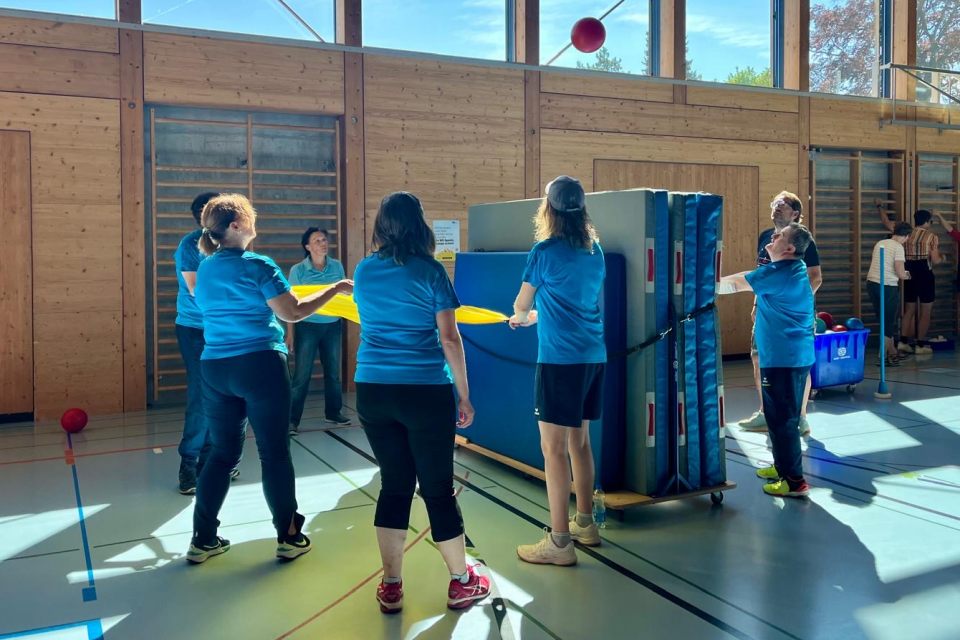 Eine Gruppe von Personen in blauen Hemden spielt in einer Halle ein Mannschaftsspiel, bei dem sie mit gelben Schlägern einen roten Ball balancieren. Sie stehen neben gestapelten Turnmatten in einer sonnenbeschienenen Sporthalle.