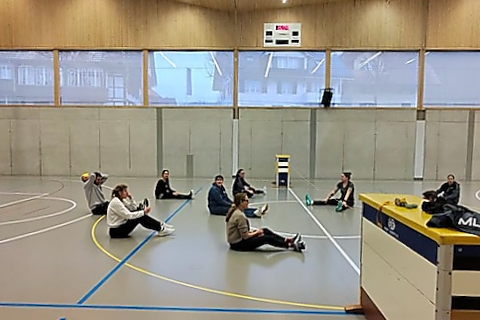 In einer großen Sporthalle sitzen mehrere Personen auf dem Boden und dehnen sich, mit etwas Abstand zueinander. Die Turnhalle hat hohe Fenster und Holzwände, und im Vordergrund sind einige Sportgeräte zu sehen.