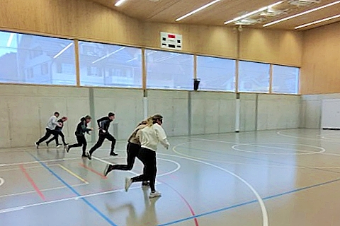 Fünf Personen rennen in einer Turnhalle mit hoher Decke und großen Fenstern; die Halle hat Markierungen auf dem Boden und eine Anzeigetafel an der Wand.