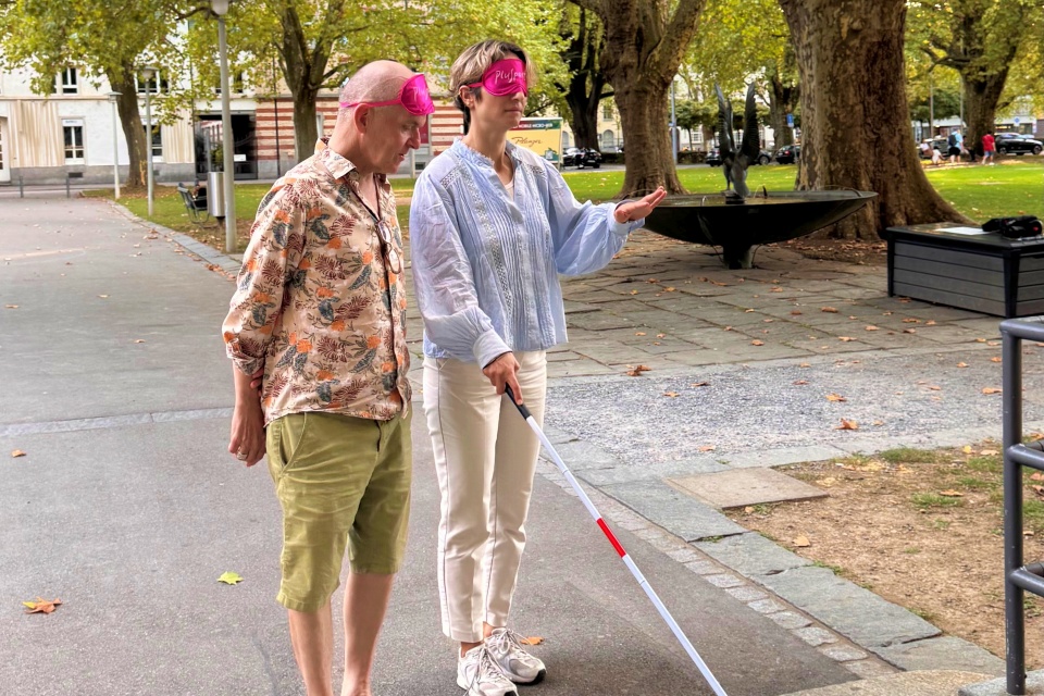 Zwei Menschen mit rosa Augenbinden gehen im Freien spazieren; einer benutzt einen weißen Blindenstock und deutet nach vorne, während der andere neben ihm hergeht. Im Hintergrund sind Bäume, Gras und Pflaster zu sehen.