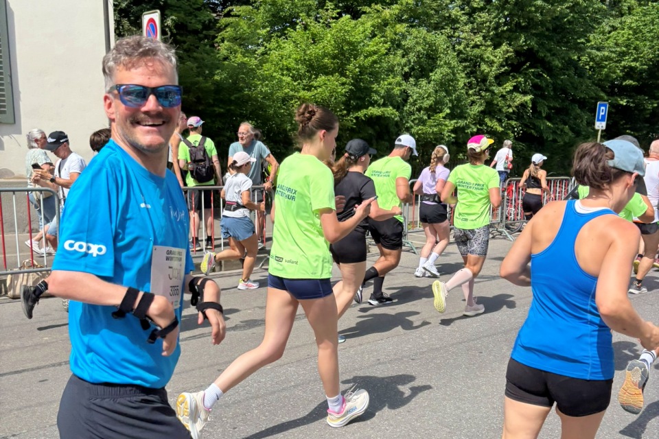 Ein lächelnder Mann in einem blauen Hemd und mit Sonnenbrille nimmt an einem Lauf teil und blickt dabei in die Kamera. Andere Läufer in grünen und blauen Hemden laufen an einem sonnigen Tag voraus, während hinter einer Metallabsperrung Bäume und Zuschauer zu sehen sind.