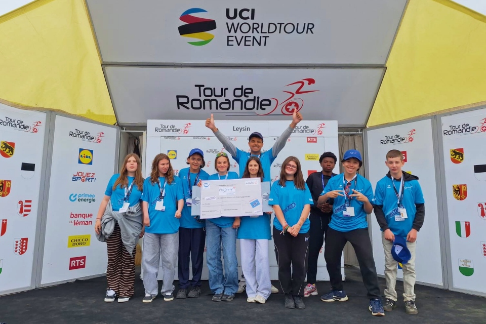 Eine Gruppe junger Menschen in blauen Hemden posiert auf der Bühne der Tour de Romandie mit einem großen Scheck in der Hand, hinter ihnen sind ein gelbes Zelt sowie die Logos der UCI WorldTour und der Sponsoren zu sehen.
