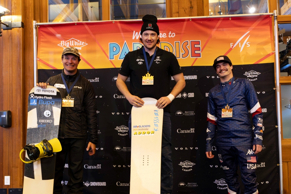 Drei Snowboarder stehen auf einem Podium mit Snowboards vor einem "Welcome to Paradise"-Banner. Der Athlet in der Mitte steht lächelnd auf dem obersten Podest, die beiden anderen stehen seitlich und halten ihre Bretter und Medaillen.