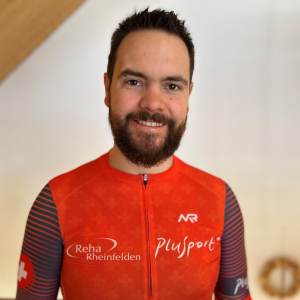 Ein Mann mit kurzen dunklen Haaren und einem Bart lächelt in die Kamera. Er trägt ein rotes Sporttop mit den Logos "Reha Rheinfelden" und "PluSport". Der Hintergrund ist in Innenräumen sanft verschwommen.