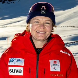 Eine lächelnde Person in einer roten Jacke mit Schweizer Paralympics- und Sponsorenlogos steht im Freien in einer verschneiten Berglandschaft, trägt eine dunkelblaue Mütze und eine Skibrille auf dem Kopf.