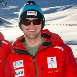 Eine Person lächelt im Freien in einer verschneiten Landschaft. Sie trägt eine marineblaue und weisse Mütze, eine dunkle Sonnenbrille und eine rote Jacke mit verschiedenen Logos, darunter Swiss Paralympic und SUVA.