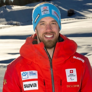 Ein Mann mit Bart lächelt im Freien in einer verschneiten Landschaft. Er trägt eine leuchtend rote Jacke mit Sponsorenlogos und ein blaues Stirnband mit der Aufschrift Swiss Paralympic. Er steht in einer sonnigen, schneebedeckten Umgebung.