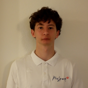 Eine junge Person mit lockigem braunem Haar steht vor einer schlichten weißen Wand und trägt ein weißes Kragenhemd mit dem Playsport-Logo darauf.