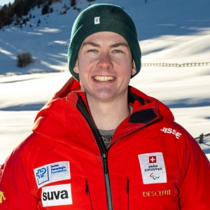 Ein junger Mensch mit grüner Mütze und roter Winterjacke mit mehreren Sponsorenlogos steht lächelnd in einer verschneiten Berglandschaft.