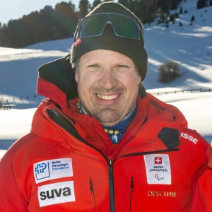 Ein Mann in einer roten Winterjacke mit mehreren Sponsorenlogos steht draußen in einer verschneiten Berglandschaft und lächelt in die Kamera. Er trägt eine schwarze Mütze, eine Skibrille und einen Pulswärmer.