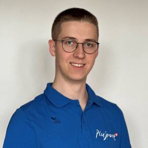 Ein junger Mann mit kurzen hellbraunen Haaren, einer runden Brille und einem schwarzen Rundhalsshirt lächelt vor einer schlichten weißen Wand in die Kamera.