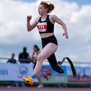 Eine Sportlerin mit einer Laufprothese nimmt an einem Rennen auf einer Freiluftbahn teil. Sie trägt ein schwarzes Sportoutfit und die Startnummer 171, mit einem entschlossenen Gesichtsausdruck und blauem Himmel im Hintergrund.