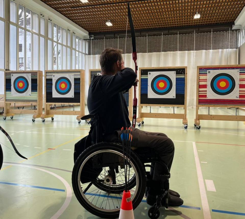 Eine Person im Rollstuhl zielt mit Pfeil und Bogen auf bunte Zielscheiben in einer Sporthalle. Mehrere Pfeile befinden sich in einem Halter, der am Rollstuhl befestigt ist. Durch große Fenster strömt natürliches Licht.