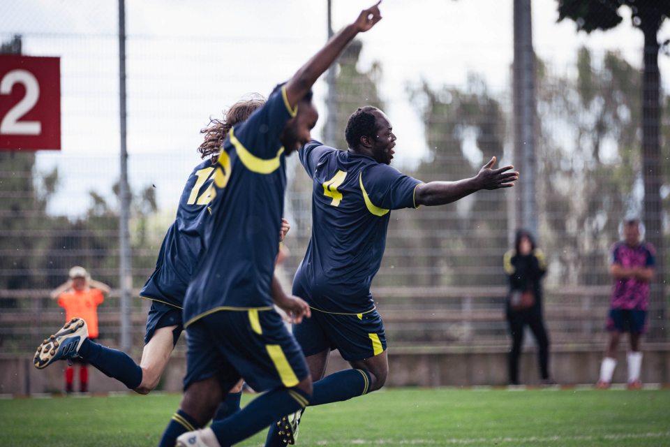 Drei Fußballspieler in blauen und gelben Trikots jubeln auf einem Feld, die Arme ausgestreckt, während sie laufen. Im Hintergrund ist eine große rote Anzeigetafel zu sehen und ein paar Leute schauen vom Spielfeldrand aus zu.