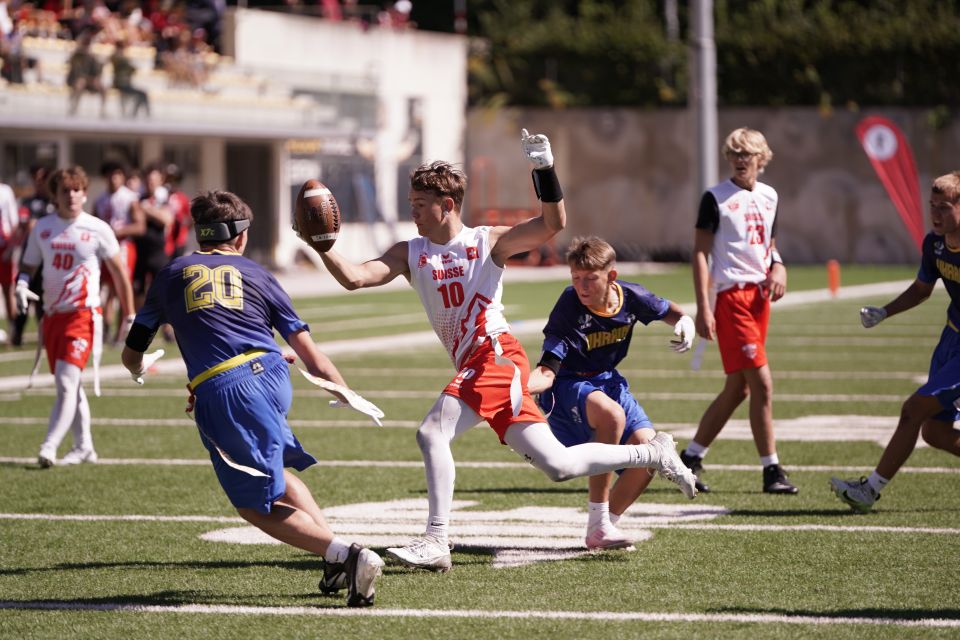 Ein Flag-Football-Spieler in einem weiß-roten Trikot rennt mit dem Ball und streckt seinen Arm aus, um zwei Verteidiger in blauen und gelben Trikots abzuwehren, während andere Spieler auf einem sonnigen Feld zusehen.