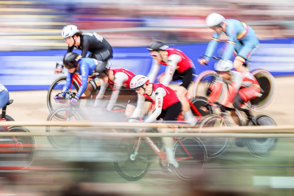 Eine Gruppe von Radfahrern in farbenfroher Rennausrüstung rast über eine Indoor-Rennstrecke und erzeugt einen Bewegungsunschärfeeffekt, während sie dicht beieinander um die Wette fahren.