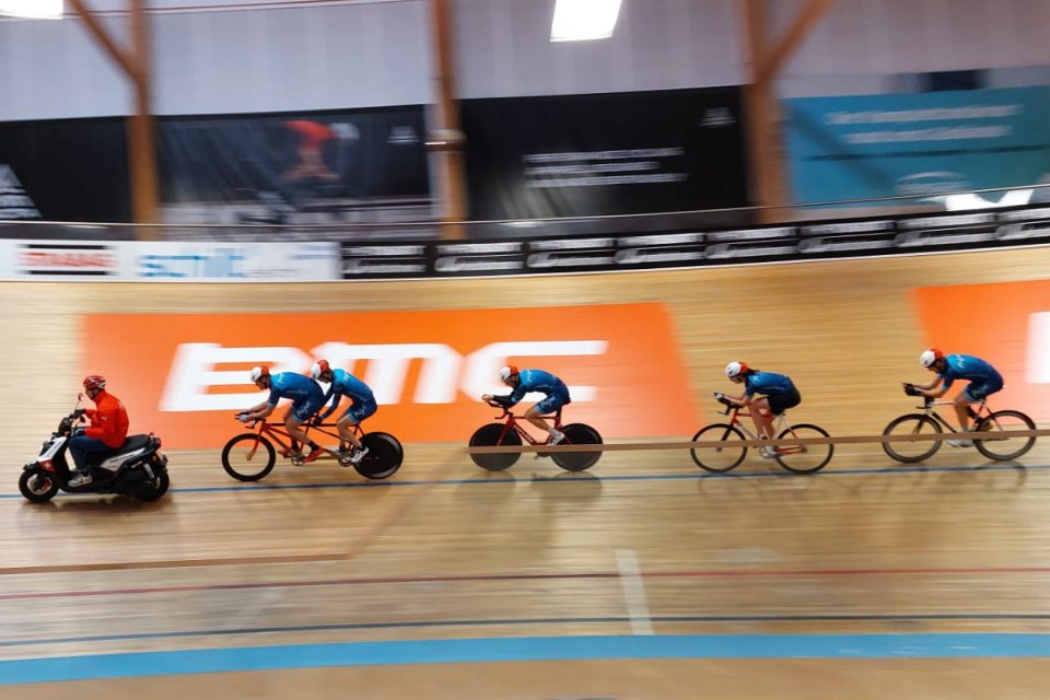 Vier Radfahrer in Blau fahren dicht hintereinander auf einer Indoor-Velodrombahn und folgen dabei einem Fahrer auf einem roten Roller. Der Hintergrund ist unscharf, was ihre Geschwindigkeit und Bewegung betont.