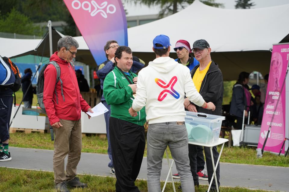 Eine Gruppe von Menschen steht im Freien unter einem weißen Zelt an einem Tisch mit Veranstaltungsmaterialien. Einige tragen bunte Jacken und Hüte, eine Person trägt einen Kapuzenpullover mit einem bunten Logo. Im Hintergrund sind Veranstaltungsbanner zu sehen.
