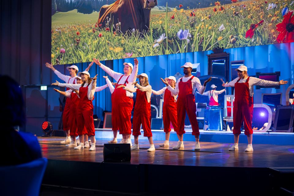 Eine Gruppe von Darstellern in roten Overalls und weißen Hemden tanzt auf der Bühne, während hinter ihnen eine große Leinwand eine Wiesenszene mit einer Kuh zeigt.