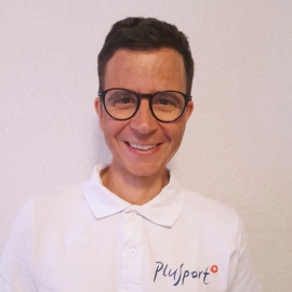Eine Person mit kurzen braunen Haaren und Brille, die lächelt; sie trägt ein weißes Hemd mit Kragen und dem PluSport-Logo und steht vor einer schlichten, hellen Wand.