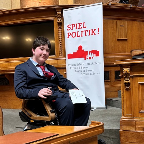 Ein junger Mann in Anzug und roter Krawatte sitzt in einem Rollstuhl und hält eine Zeitung in der Hand vor einem „Spiel Politik!“-Banner in einem formellen, holzgetäfelten Raum mit Sitzen und Schreibtischen.
