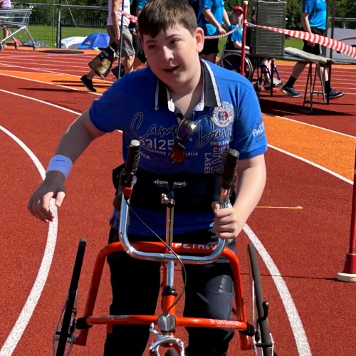 Ein Junge in einem blauen Hemd fährt während einer Outdoor-Veranstaltung mit einem adaptiven Dreirad auf einer Laufstrecke. Im Hintergrund sind weitere Personen und Veranstaltungsausrüstung zu sehen.