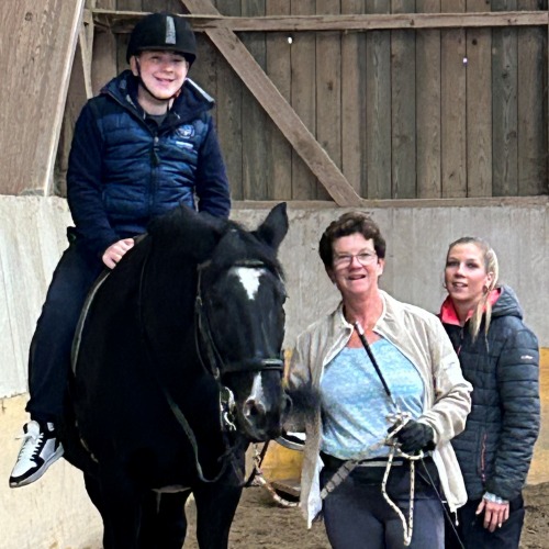 Ein lächelnder junger Mensch mit Helm reitet in der Halle auf einem schwarzen Pferd. Neben dem Pferd stehen zwei Frauen, die alle fröhlich und entspannt wirken.