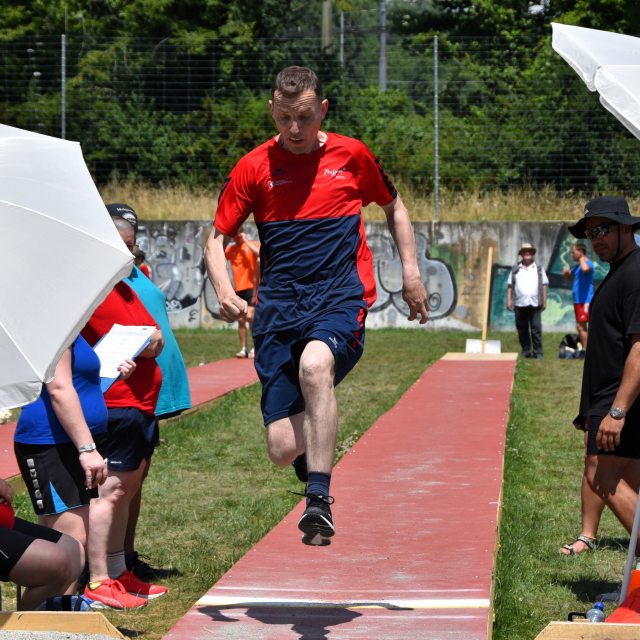 Ein Mann in roter und blauer Sportkleidung befindet sich in der Luft, während er einen Weitsprung auf einer Freiluftbahn ausführt, während im Hintergrund Zuschauer und Offizielle zusehen.
