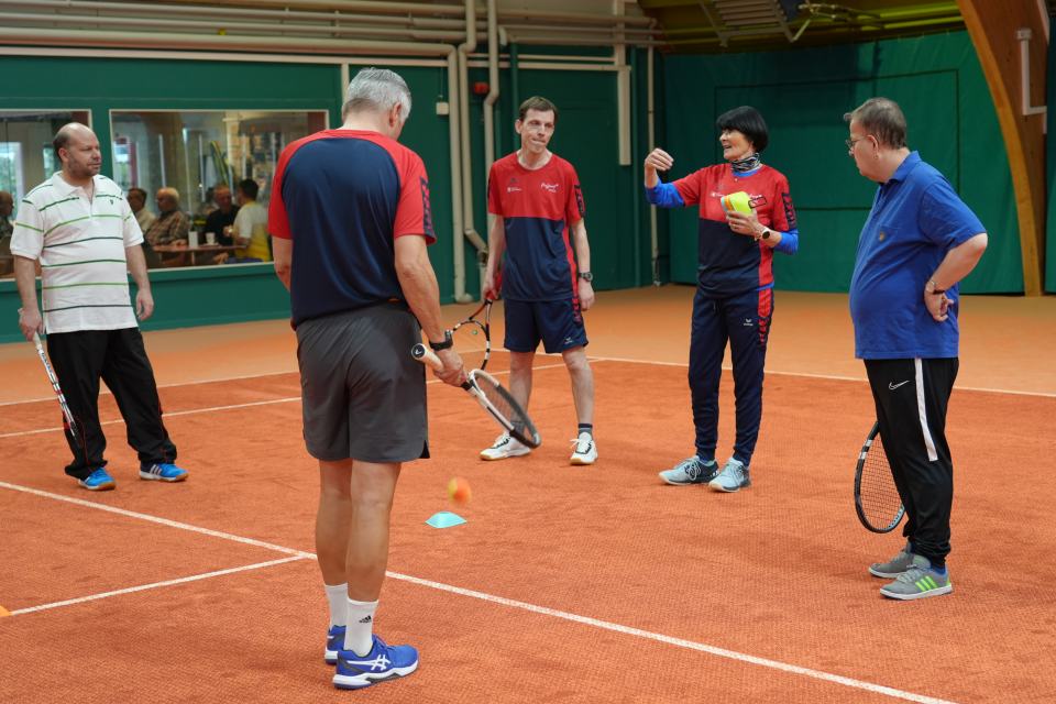 Fünf Erwachsene stehen auf einem Hallen-Tennisplatz, halten Schläger in der Hand und hören einem Trainer zu. Sie scheinen sich zu unterhalten oder zu unterrichten, umgeben von der Tennisausrüstung auf dem orangefarbenen Platz.