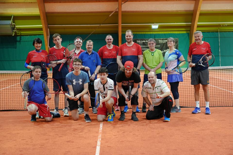 Eine Gruppe von fünfzehn Personen, die Tennisschläger in der Hand halten und lächeln, posiert auf einem überdachten Tennisplatz vor dem Netz. Die meisten tragen Sportkleidung, und der Platz hat eine orangefarbene Sandoberfläche.