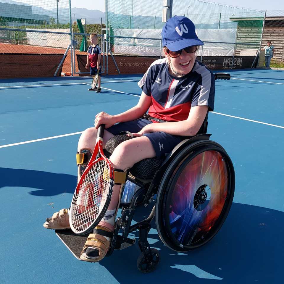 Ein lächelnder Tennisspieler im Rollstuhl hält einen Schläger auf einem blauen Tennisplatz im Freien. Er trägt eine Kappe, Sportuniform und eine Sonnenbrille. Im Hintergrund sind ein weiterer Spieler und Tennisnetze zu sehen.